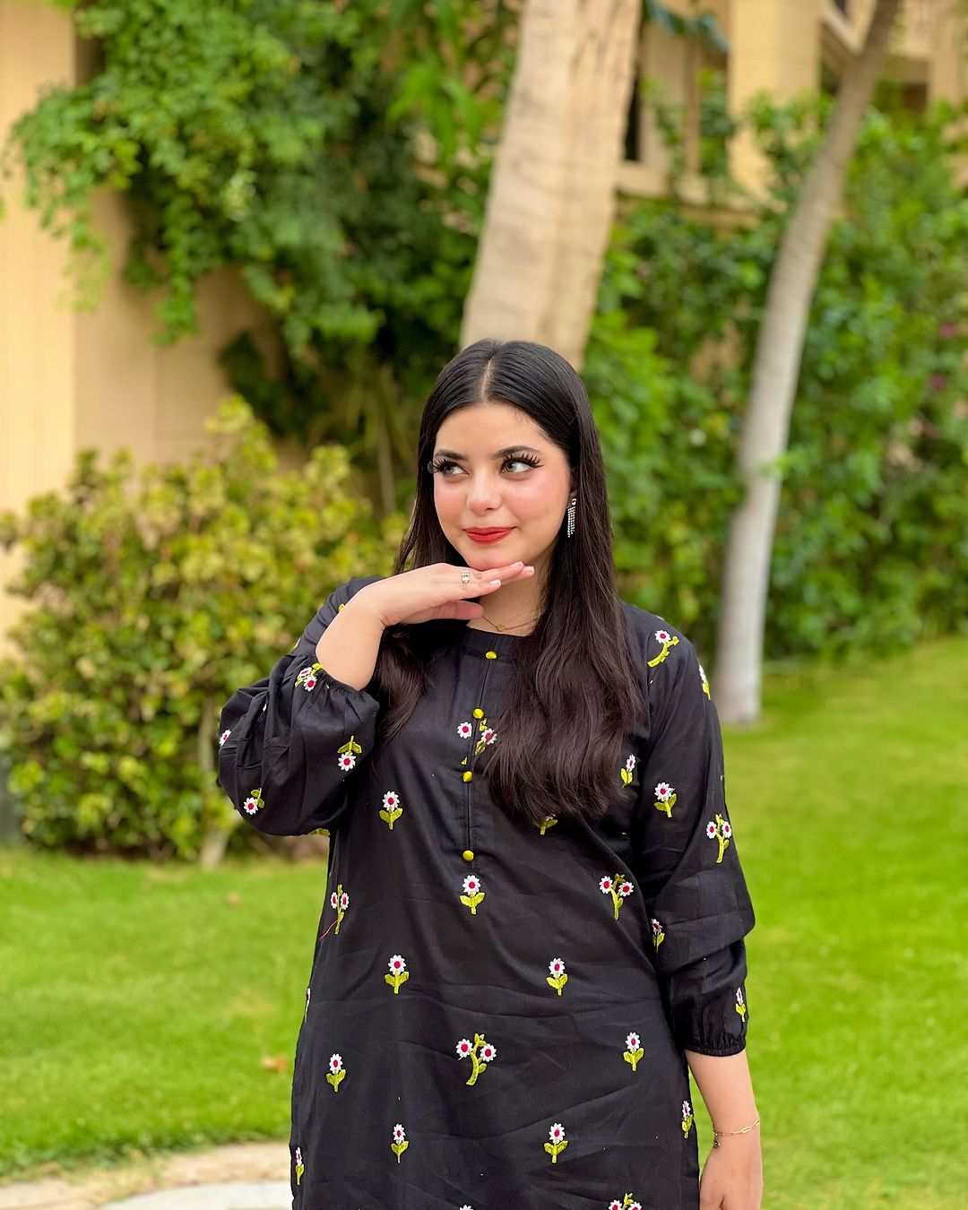 Woman in a black dress with floral patterns standing outdoors with greenery in the background