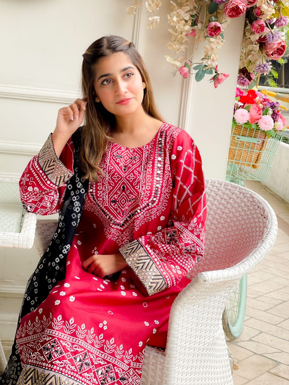 Woman in a red and white patterned outfit sitting on a white chair with floral decorations in the background.