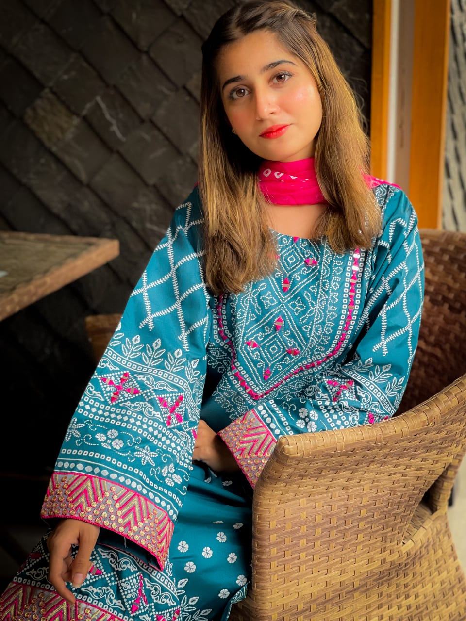 Woman wearing a blue and pink traditional outfit sitting on a woven chair.