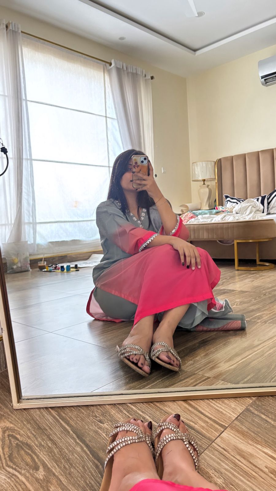 Woman taking a mirror selfie in a bedroom with a bed and lamp in the background.