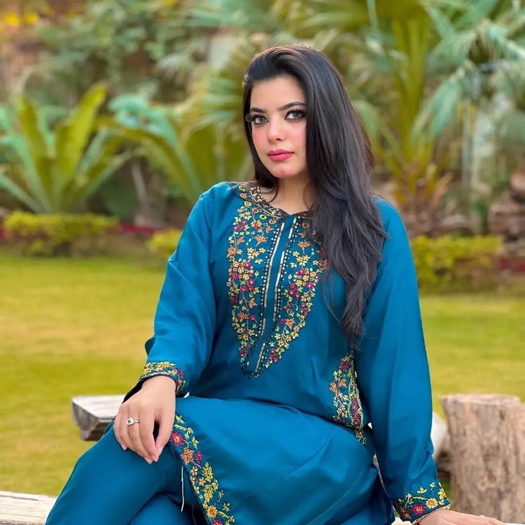 Woman in a blue embroidered outfit sitting outdoors with greenery in the background