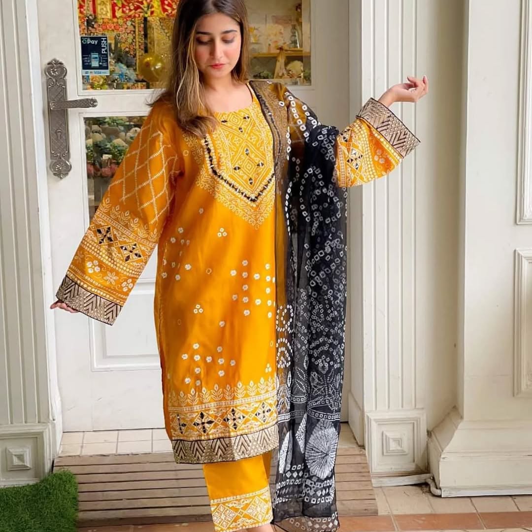 Woman holding a yellow embroidered dress with a black and white patterned dupatta in a room.