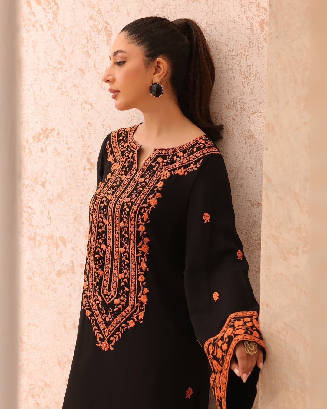 Woman wearing a black and orange embroidered traditional outfit against a beige wall.