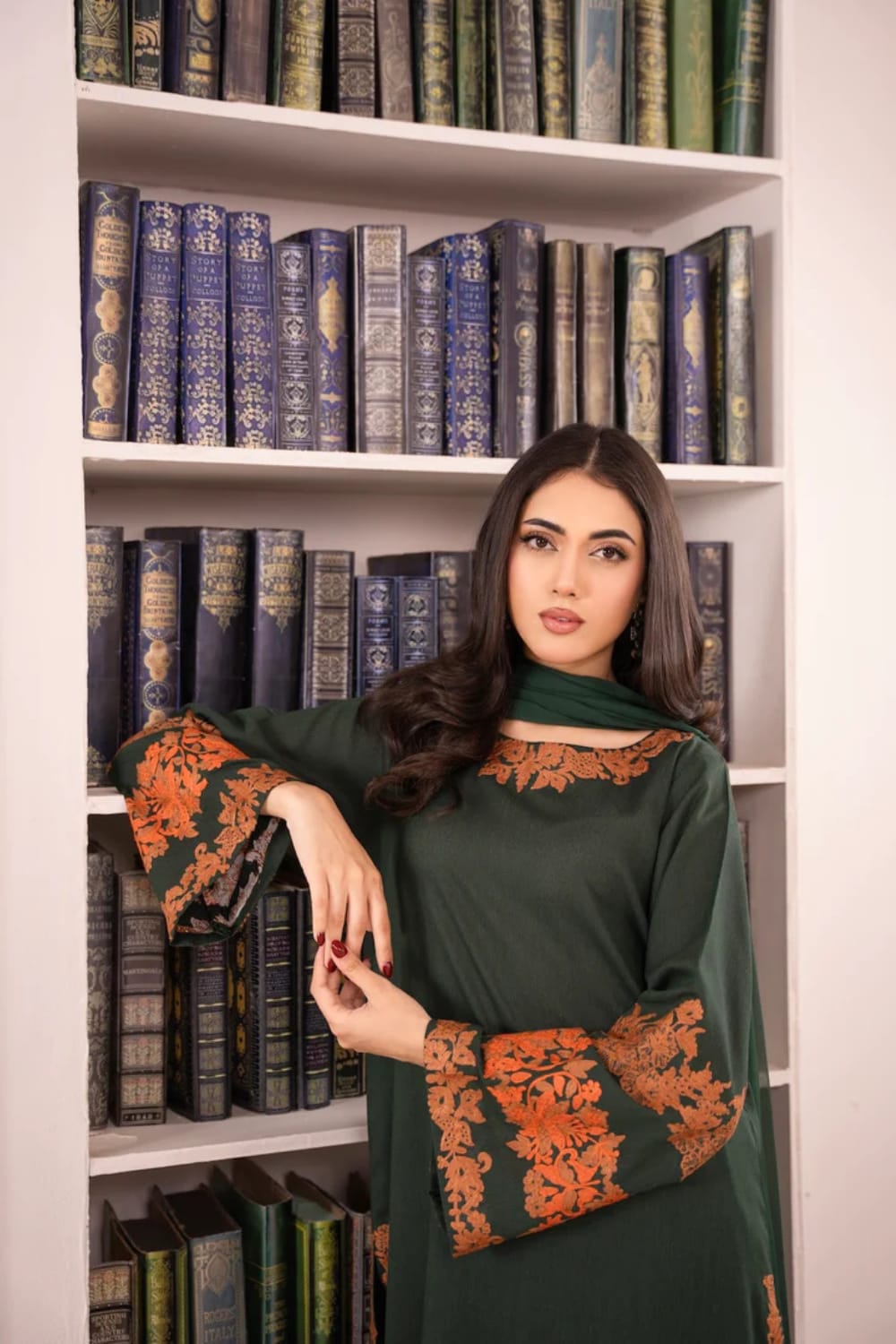 Woman in a green dress with orange embroidery standing in front of a bookshelf filled with books.